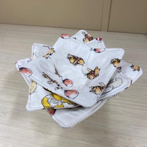 May include: Stack of three white fabric bowl cozies with a cartoon bear print. The cozies are square with a star-like shape and a quilted texture. The print features the bear, balloons, and other cartoon elements.