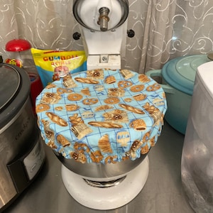 May include: A white KitchenAid stand mixer with a blue and tan fabric bowl cover featuring a pattern of baked goods. The cover is gathered at the edge and fits over the mixing bowl. Other kitchen items are visible in the background.