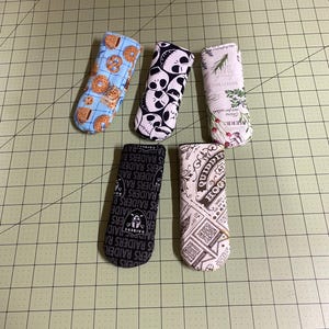 May include: Five fabric glasses cases with various patterns. One case has a blue background with pretzels and donuts. Another features black skulls on a white background. Other cases have herb, Raiders, and Hogwarts designs.