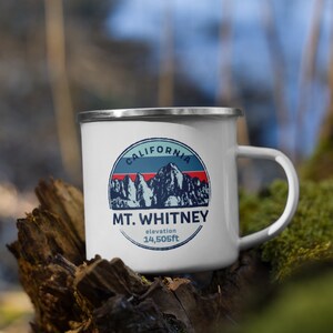 Può includere: Tazza smaltata bianca con bordo e manico argentati. La tazza presenta una grafica della catena montuosa del Monte Whitney con la scritta "CALIFORNIA" e "MT. WHITNEY altitudine 4.421m" in un design circolare. La tazza è appoggiata su un pezzo di legno.
