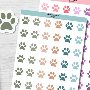 May include: A sheet of paw print stickers with various colors, including green, brown, teal, and blue. The stickers are arranged in rows and columns on a white background. The text "Jude Anne Designs" is printed at the top of the sheet.