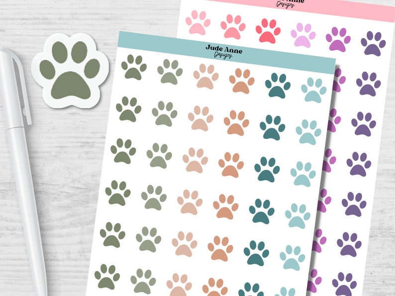 May include: A sheet of paw print stickers with various colors, including green, brown, teal, and blue. The stickers are arranged in rows and columns on a white background. The text "Jude Anne Designs" is printed at the top of the sheet.