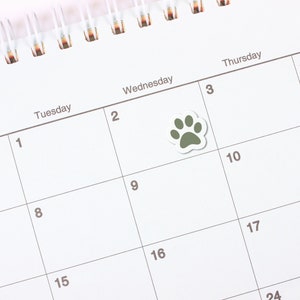 May include: A white calendar with a green paw print sticker on the third square. The calendar has the days of the week labeled at the top and numbers for each day. A white pen is lying on the calendar.