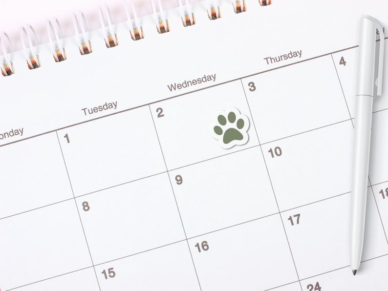 May include: A white calendar with a green paw print sticker on the third square. The calendar has the days of the week labeled at the top and numbers for each day. A white pen is lying on the calendar.