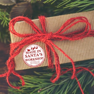 Puede incluir: Una caja de regalo marr&oacute;n atada con cuerda roja y un lazo rojo. Una pegatina blanca con el texto "MADE IN SANTA'S WORKSHOP" est&aacute; en la caja.