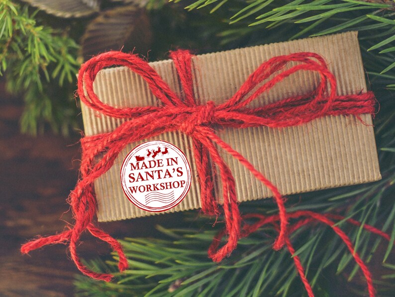 Puede incluir: Una caja de regalo marr&oacute;n atada con cuerda roja y un lazo rojo. Una pegatina blanca con el texto "MADE IN SANTA'S WORKSHOP" est&aacute; en la caja.