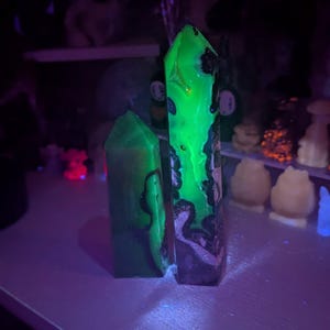 Puede incluir: Dos torres de cristal hexagonales con un patrón jaspeado en verde oscuro y negro. Bajo luz ultravioleta, la torre más alta muestra una luminiscencia verde neón vibrante en su superficie, mientras que la torre más corta permanece oscura.