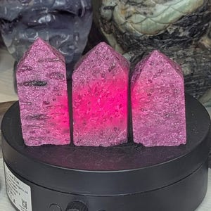 Op de afbeelding: Drie levendige magentakleurige kristalpunten met een ruw, getextureerd oppervlak en puntige toppen. Deze stenen torens staan naast elkaar op een zwarte ronde draaibare displaystandaard met een bedieningsknop en een wit productlabel.