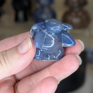 May include: A small, polished blue stone carving shaped like a turtle with a smooth, glossy finish. The figurine features a rounded shell, a distinct head, and four flippers, showcasing a deep, translucent blue color with subtle internal variations.