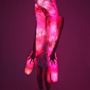 Peut inclure: Ensemble rose en fausse fourrure avec capuche et écharpe à lumières LED intégrées. La capuche couvre la tête et se prolonge en une longue écharpe, créant un effet lumineux. La tenue est complétée par une jupe assortie et des bottines à plateforme, le tout illuminé de rose.