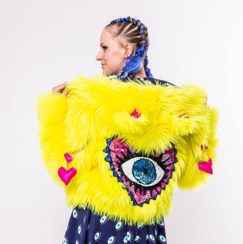 May include: A bright yellow faux fur jacket with a large sequined eye and heart design on the front. The jacket has pink hearts and a silver chain hanging from the collar.