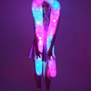 May include: A white faux fur hood with cat ears and matching scarf, illuminated with LED lights in shades of pink, blue, and white. The outfit is worn with a white dress and platform boots, creating a vibrant, eye-catching look.