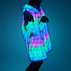 May include: A sleeveless vest made of long-pile faux fur that is illuminated with integrated LED lights. The fur displays a vibrant, multi-colored glow, shifting between shades of bright neon blue, vivid pink, and electric green throughout the fabric.