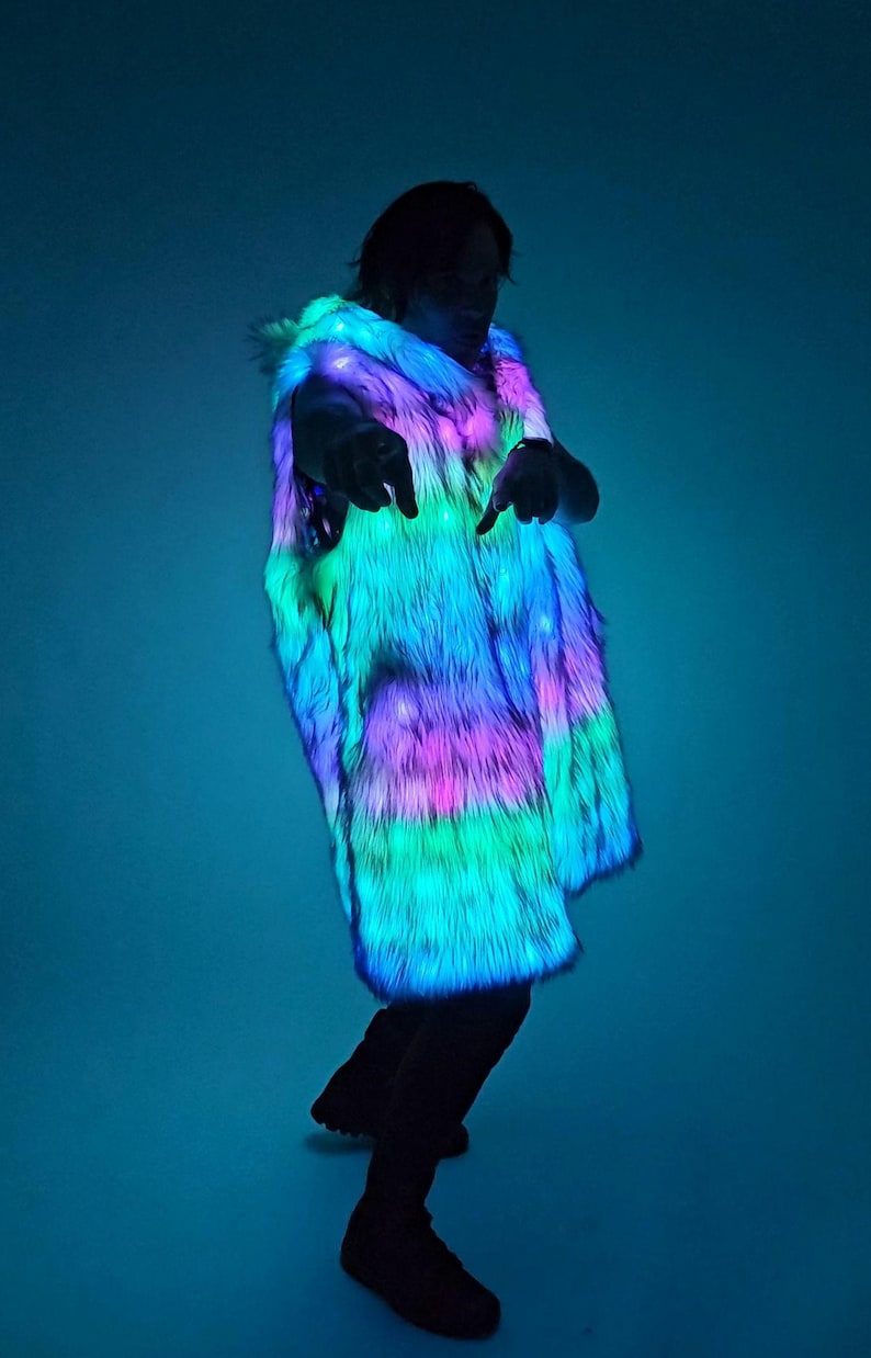 May include: A sleeveless vest made of long-pile faux fur that is illuminated with integrated LED lights. The fur displays a vibrant, multi-colored glow, shifting between shades of bright neon blue, vivid pink, and electric green throughout the fabric.