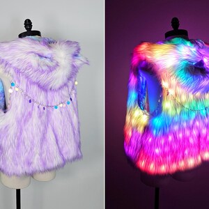 May include: A sleeveless faux fur vest with a hood, featuring a light purple and white color palette. A silver-toned chain with small, circular, iridescent sequins is draped across the back. The vest is equipped with integrated LED lights.