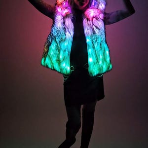 May include: A sleeveless faux fur vest featuring an integrated hood with pointed ears. The garment is illuminated by internal LED lights that emit a vibrant gradient of green, pink, and blue hues throughout the textured, shaggy white fur material.