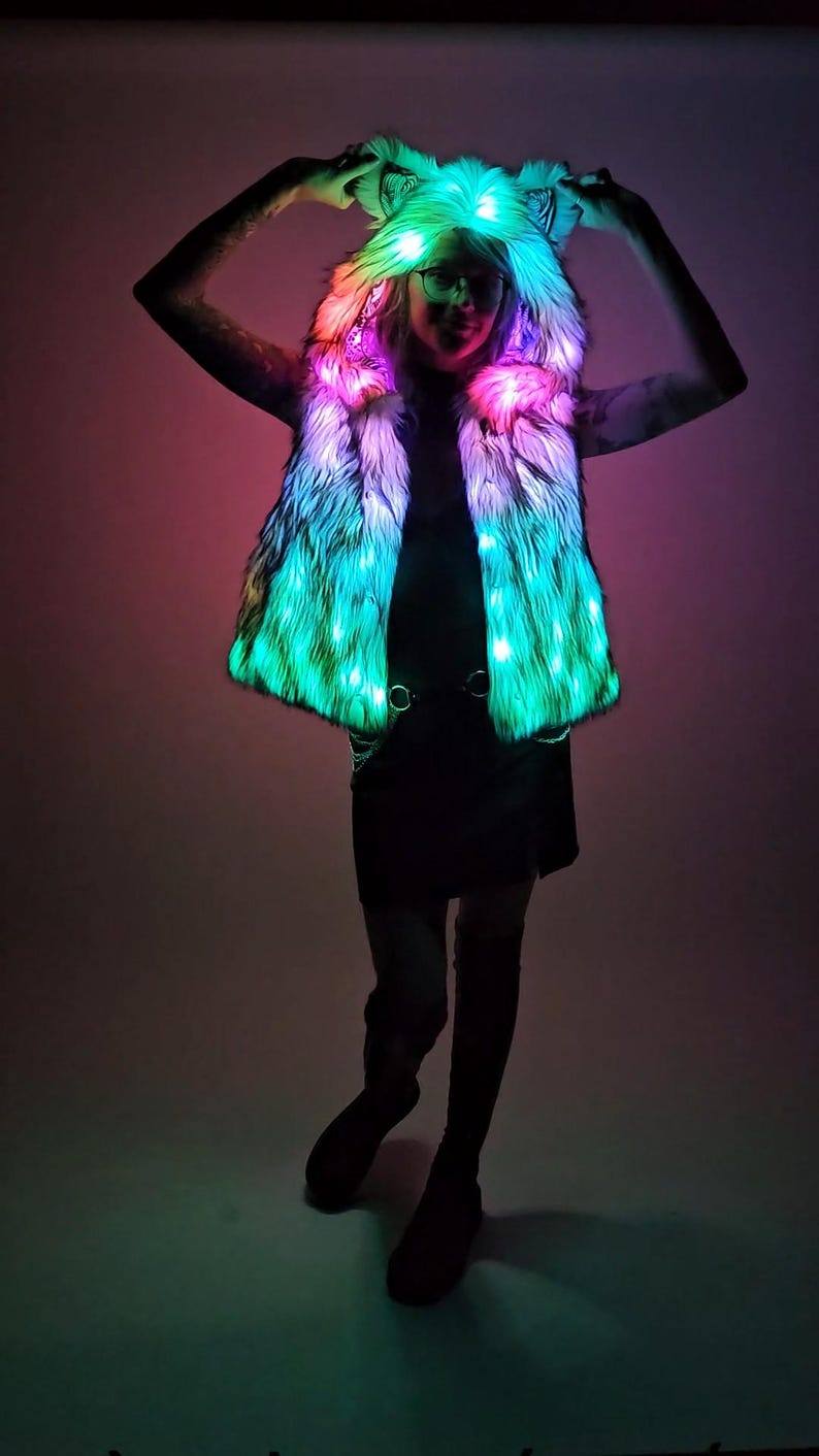 May include: A sleeveless faux fur vest featuring an integrated hood with pointed ears. The garment is illuminated by internal LED lights that emit a vibrant gradient of green, pink, and blue hues throughout the textured, shaggy white fur material.