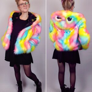 May include: A rainbow-colored faux fur jacket with a hood featuring a sequined eye design. The jacket is cropped and has a relaxed fit.