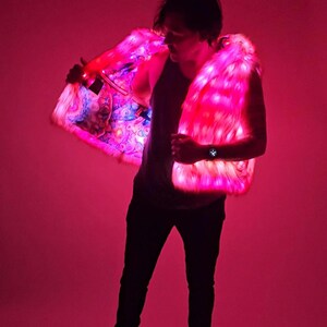 May include: A sleeveless vest made of faux fur that is illuminated with bright pink LED lights. The interior lining of the vest features a colorful, multi-patterned graphic print. The garment is worn over a dark tank top and dark trousers.