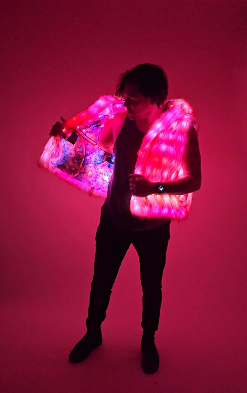 May include: A sleeveless vest made of faux fur that is illuminated with bright pink LED lights. The interior lining of the vest features a colorful, multi-patterned graphic print. The garment is worn over a dark tank top and dark trousers.