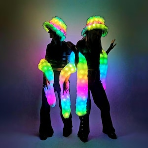 Puede incluir: Dos personas con sombreros de colores brillantes y peludos y brazaletes a juego que se iluminan. Los sombreros y los brazaletes son de colores arcoíris y tienen una textura peluda.