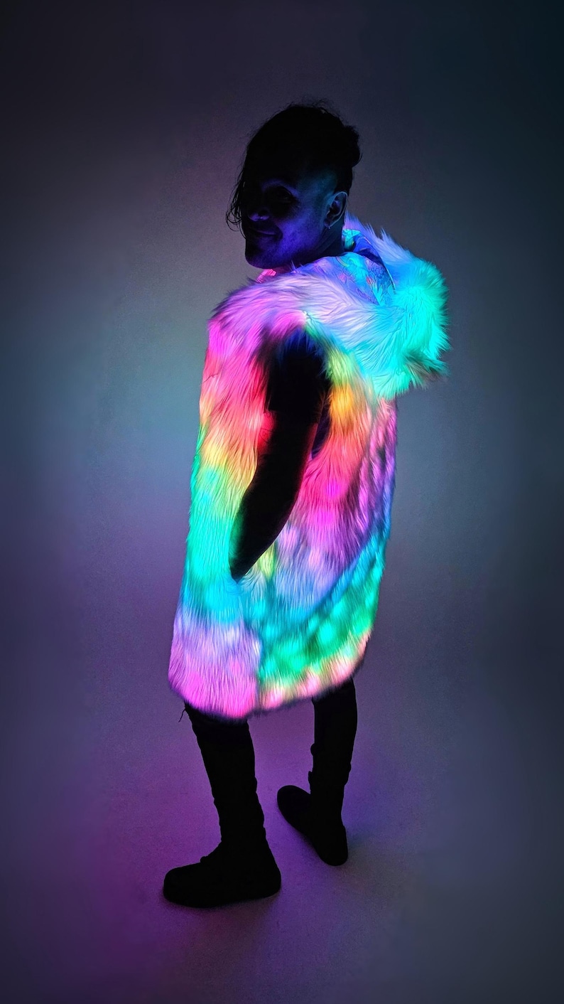May include: A sleeveless hooded vest made of long-pile faux fur that is illuminated with vibrant, multi-colored LED lights. The vest features a gradient of glowing pink, blue, and yellow hues throughout the fur, creating a bright, light-up effect.