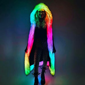Puede incluir: Una persona lleva un abrigo de piel sintética arcoíris con capucha. El abrigo está iluminado con luces LED, creando un efecto vibrante y colorido.