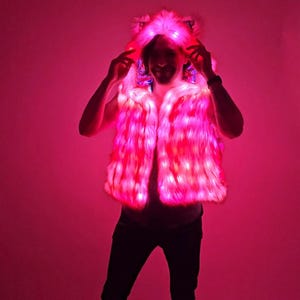 May include: A sleeveless faux fur vest featuring an integrated hood with animal-like ears. The garment is illuminated by bright pink LED lights embedded throughout the plush material, creating a vibrant glow across the entire surface of the vest.