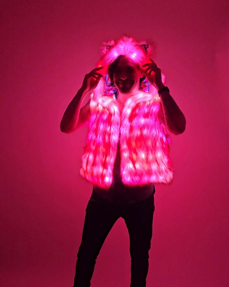 May include: A sleeveless faux fur vest featuring an integrated hood with animal-like ears. The garment is illuminated by bright pink LED lights embedded throughout the plush material, creating a vibrant glow across the entire surface of the vest.
