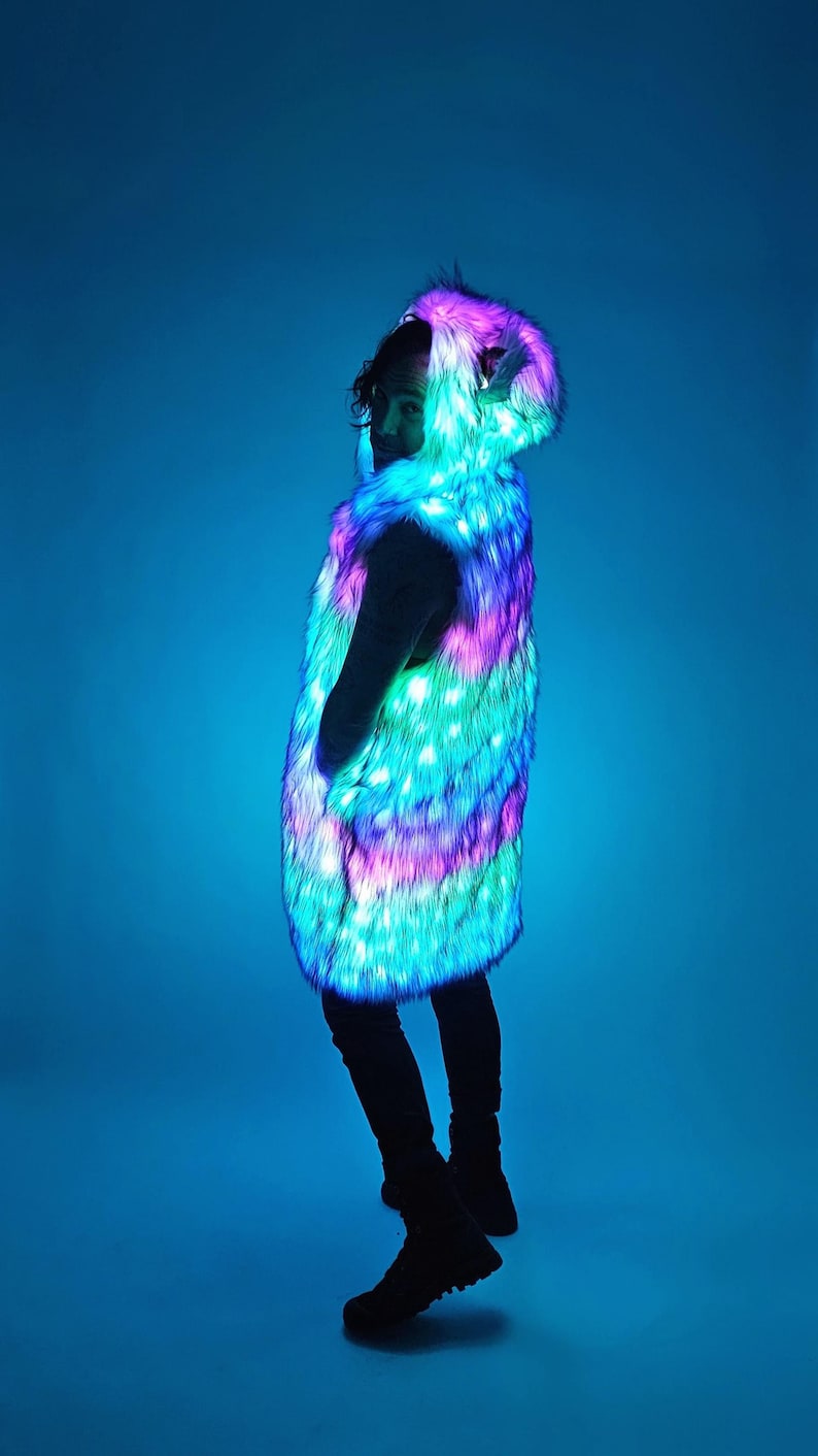 May include: A sleeveless faux fur vest featuring an integrated LED lighting system that emits vibrant pink, purple, and cyan light. The vest includes a hood with small ears and a long, straight-cut silhouette that reaches the mid-thigh area.