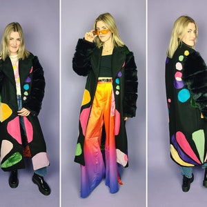 May include: A black long coat with colorful abstract shapes and fur trim on the sleeves. The coat is worn over a rainbow ombre pants.