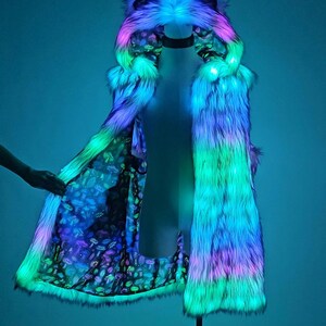 May include: A long, sleeveless faux fur coat featuring an integrated hood with ears, illuminated by vibrant blue, green, and pink LED lights. The interior lining displays a dark fabric pattern filled with colorful, detailed mushroom illustrations.