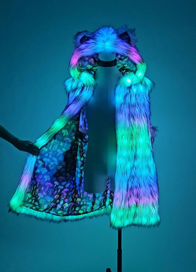 May include: A long, sleeveless faux fur coat featuring an integrated hood with ears, illuminated by vibrant blue, green, and pink LED lights. The interior lining displays a dark fabric pattern filled with colorful, detailed mushroom illustrations.