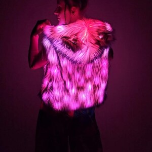 May include: A sleeveless hooded vest made of faux fur material that is illuminated with bright pink LED lights. The vest features a textured, shaggy appearance and includes integrated lighting elements throughout the fabric, creating a vibrant glow.