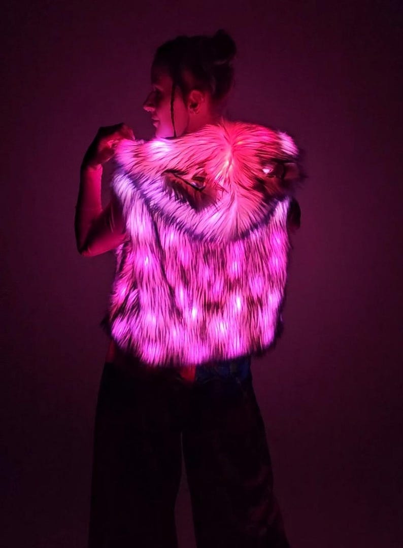 May include: A sleeveless hooded vest made of faux fur material that is illuminated with bright pink LED lights. The vest features a textured, shaggy appearance and includes integrated lighting elements throughout the fabric, creating a vibrant glow.