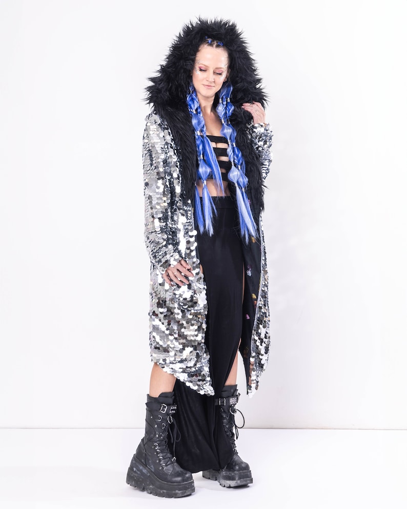 May include: A silver sequined coat with a black faux fur hood and lining. The coat has a long, flowing silhouette and is worn over a black skirt.