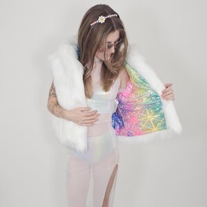 May include: A white faux fur vest featuring a vibrant, multicolored holographic interior lining with a geometric star pattern. The vest is worn over a sheer, iridescent white bodysuit with high side slits and a matching daisy headband.