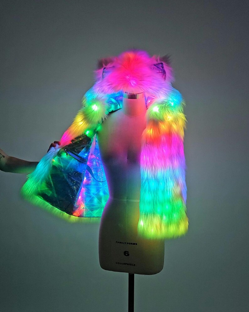 May include: A sleeveless faux fur vest with an integrated hood featuring animal-like ears. The garment is illuminated by vibrant, multi-colored LED lights that transition through shades of pink, blue, green, and yellow across the plush texture.