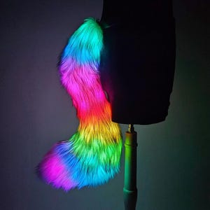 Op de afbeelding: Een levendige, regenboogkleurige imitatiebontstaart, weergegeven op een zwarte mannequin. De staart gaat over in tinten roze, oranje, geel, groen en blauw. De verlichting creëert een gloeiend effect, dat de zachte textuur benadrukt.