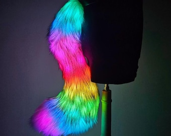 May include: A vibrant, rainbow-colored faux fur tail, displayed on a black mannequin. The tail transitions through shades of pink, orange, yellow, green, and blue. The lighting creates a glowing effect, highlighting the fluffy texture.