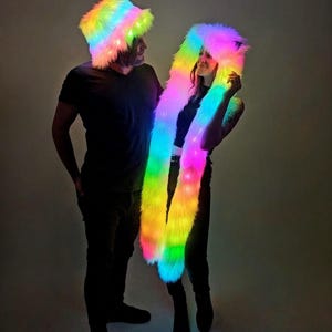 Peut inclure: Un couple portant des accessoires assortis en fausse fourrure arc-en-ciel et lumineux. L'un porte un chapeau de pêcheur, l'autre une capuche avec de longues écharpes fluides. Les accessoires brillent de couleurs vives, dont le rose, le jaune, le vert et le bleu.