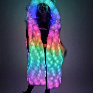 May include: A long, sleeveless faux fur coat featuring an integrated hood with pointed ears. The garment is illuminated with vibrant, multi-colored LED lights that shift between shades of pink, blue, and green throughout the textured fur material.