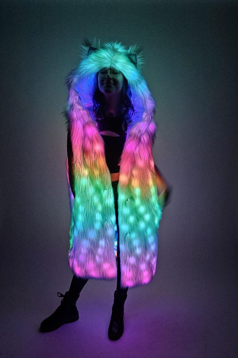 May include: A long, sleeveless faux fur coat featuring an integrated hood with pointed ears. The garment is illuminated with vibrant, multi-colored LED lights that shift between shades of pink, blue, and green throughout the textured fur material.