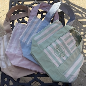 May include: A collection of four small canvas tote bags featuring vertical stripes in pastel shades of green, blue, pink, and beige. Each bag has two handles and a solid-colored base. The front-most green bag displays the Trader Joe's logo.