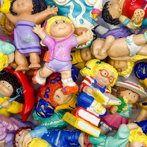 Op de afbeelding: Een verzameling kleurrijke plastic figuurtjes van kinderen, sommige met boeken, andere met paraplu's en sommige met andere accessoires. De figuurtjes staan allemaal in verschillende poses en uitdrukkingen.