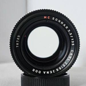 May include: A black Carl Zeiss Jena DDR MC Sonnar 3.5/135 camera lens with a ridged focus ring. The lens has the number 78725 printed on it. The lens is sitting on a black lens cap.