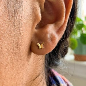 May include: A gold butterfly stud earring is shown close up. The earring is small and sits on the earlobe. The butterfly design is detailed and the earring has a shiny finish. The background is blurred.