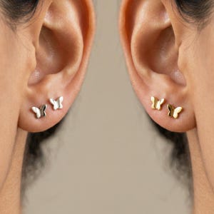 May include: Two ears each displaying butterfly stud earrings. One ear has silver butterfly studs, the other gold. The earrings are small and delicate, adding a subtle touch of elegance. The earrings are a simple, yet stylish accessory.