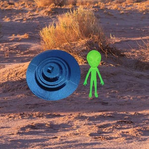 May include: A blue, circular object with a spiral design and a small green alien figurine are set against a desert landscape. The alien has a large head and slender limbs. The background features dry brush and a hazy horizon.