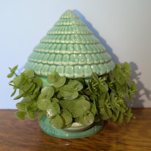 May include: A light green ceramic decorative piece with a conical top, resembling a stylized tree. The piece is adorned with a textured pattern and filled with green foliage. The item is placed on a wooden surface.